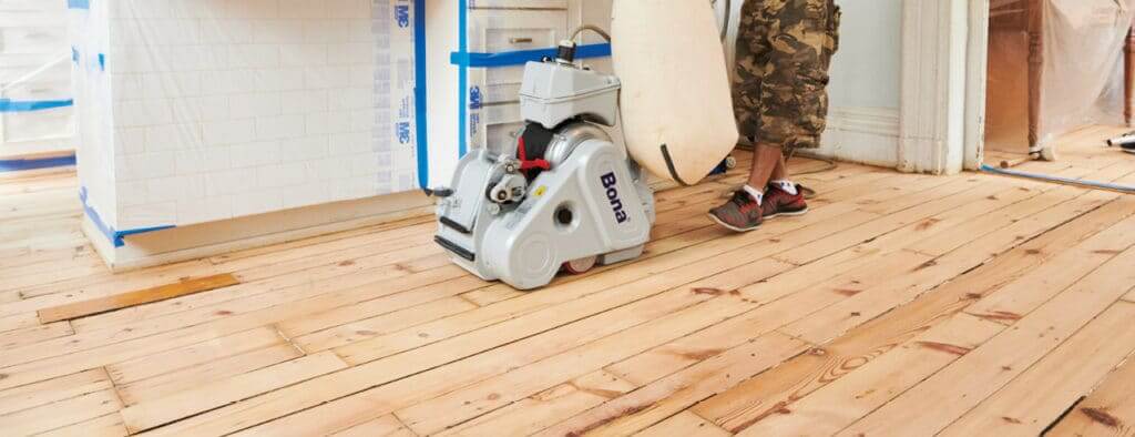 A man from Mr. Sander® is sanding a pine floor in a living room in the UK using a Bona Belt UX (AMK173100) belt sander. The machine features a 2.2 kW motor, 230 V voltage, 50 Hz frequency, 200x750 mm sanding belt size, 200 mm drum width, and weighs 85 kg.
