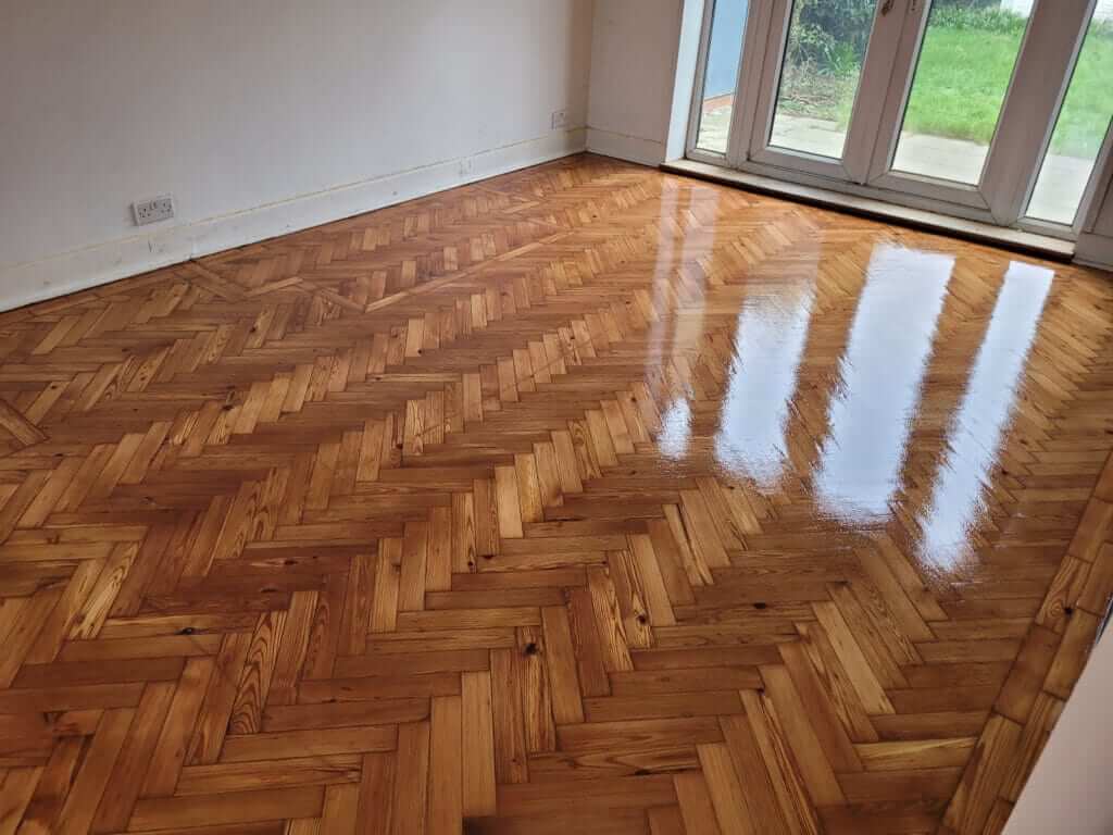 A beautifully restored herringbone wooden floor in a living room with French doors leading to a garden in South Woodford, UK. The high-gloss finish on the floor reflects the natural light, showcasing the intricate wood grain and pattern.