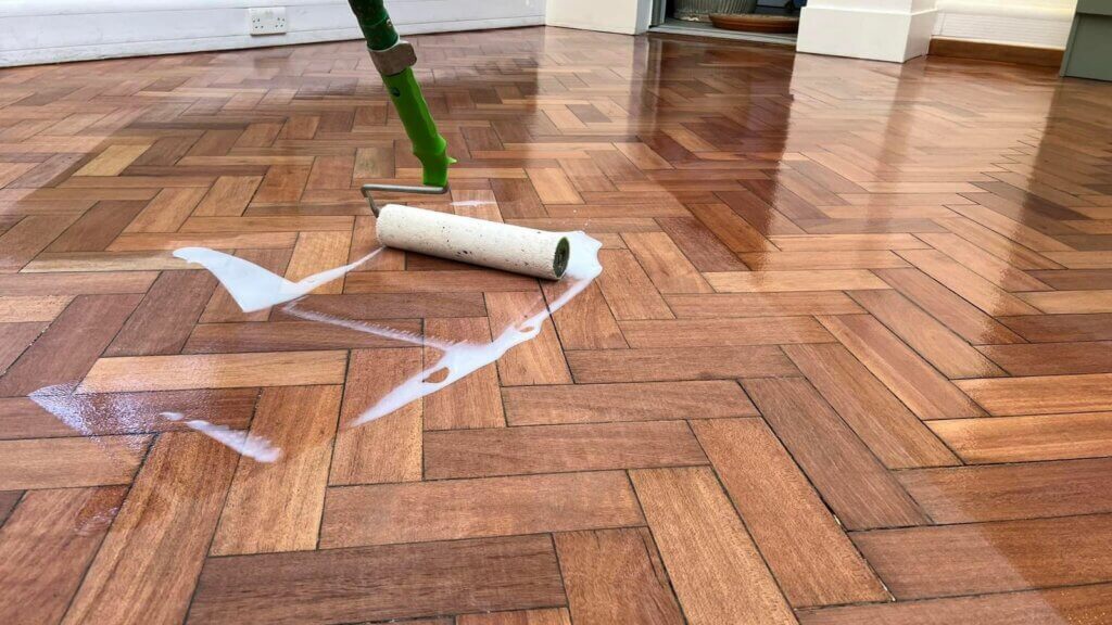 A close-up of a roller being used to apply a protective finish to a beautifully restored parquet floor. The image shows the smooth and glossy surface achieved through the final application, highlighting the meticulous care taken in parquet flooring restoration.