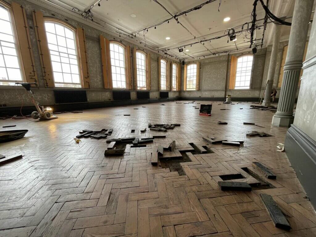A large room with high ceilings and tall windows undergoing parquet flooring restoration by Mr Sander®. The floor shows removed and damaged parquet blocks scattered across the space, indicating the initial phase of restoration. This image highlights the extensive and detailed work involved in restoring parquet flooring.