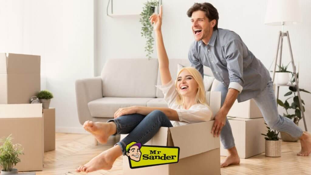 A cheerful couple enjoying their new home, with the woman sitting in a cardboard box and the man pushing her. The room features newly refinished hardwood floors and is filled with natural light. Moving boxes and potted plants are visible in the background, along with a cozy sofa. A "Mr Sander®" logo is visible at the bottom of the image.