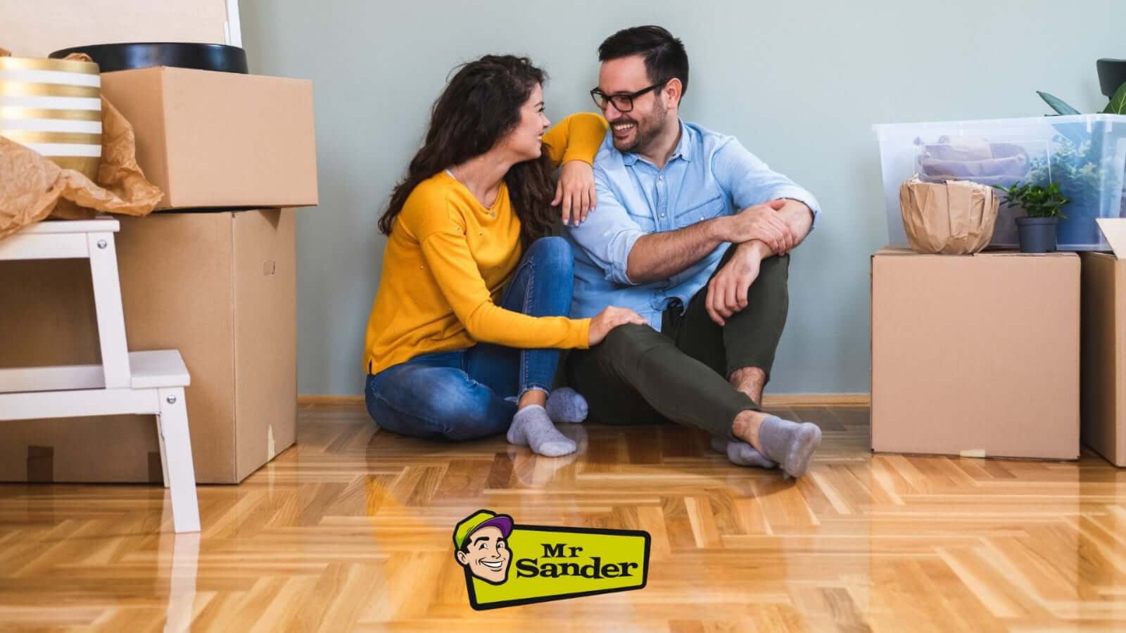 A happy couple sitting on a beautifully refinished wooden floor surrounded by moving boxes. The floor has a shiny, polished finish, showcasing the high-quality sanding and refinishing work done by Mr Sander®. The Mr Sander® logo is visible at the bottom of the image.