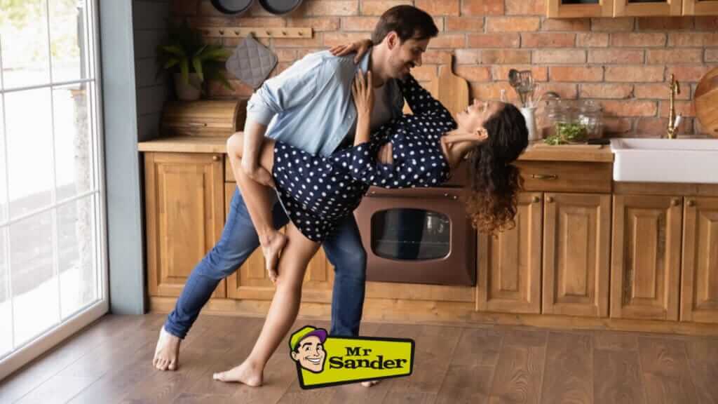 A couple dancing in their kitchen on beautifully refinished wooden floors. The kitchen has wooden cabinets, a brick backsplash, and a large window letting in natural light. A Mr Sander® logo is prominently displayed at the bottom.