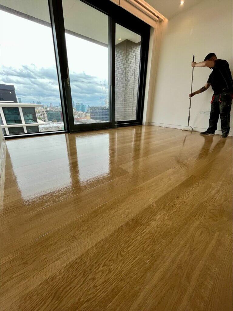 A professional from Mr Sander® applying a clear finish to a newly sanded wooden floor in a high-rise apartment with large windows overlooking a cityscape. The floor has a glossy finish, reflecting the natural light from the windows.