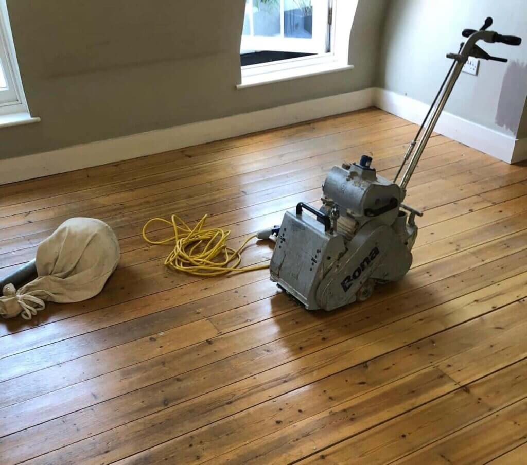 Mr Sander® is expertly sanding an oak hardwood floor in a living room in Lower Edmonton, UK, using a Bona Belt (AMO110000.3) belt sander. The Bona Belt has an effect of 2.2 kW, voltage of 230 V, frequency of 50 Hz, and dimensions of 250x750 mm. It features a drum width of 200 & 250 mm, a sanding belt size of 200 x 750 mm or 250 x 750 mm, and a total weight of 74 kg.
