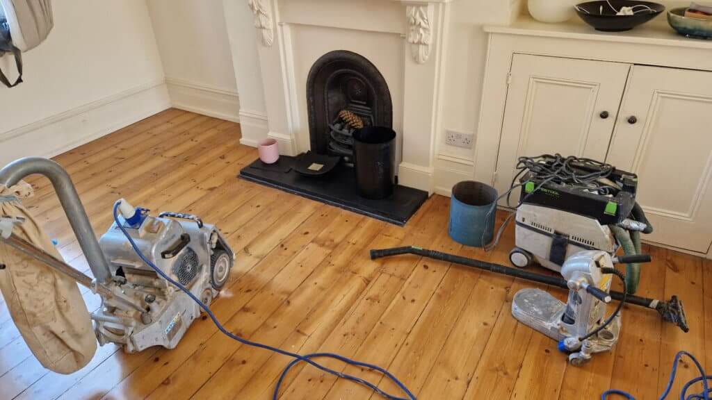 A professional from Mr Sander® is sanding a wooden floor in a living room in the UK using advanced sanding machines. He is operating a Bona Belt (AMO110000.3) with 2.2 kW motor power, 230 V voltage, and a drum width of 200 & 250 mm, complemented by a Bona Edge UX (AMO230600) edger.