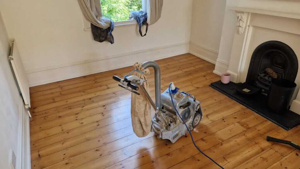 A professional from Mr Sander® is sanding a pine floor in, UK living room using a Bona Belt sander (AMO110000.3). The machine features a 2.2 kW motor, 230 V voltage, 50 Hz frequency, and a sanding belt size of 200 x 750 mm or 250 x 750 mm. The total weight is 74 kg.