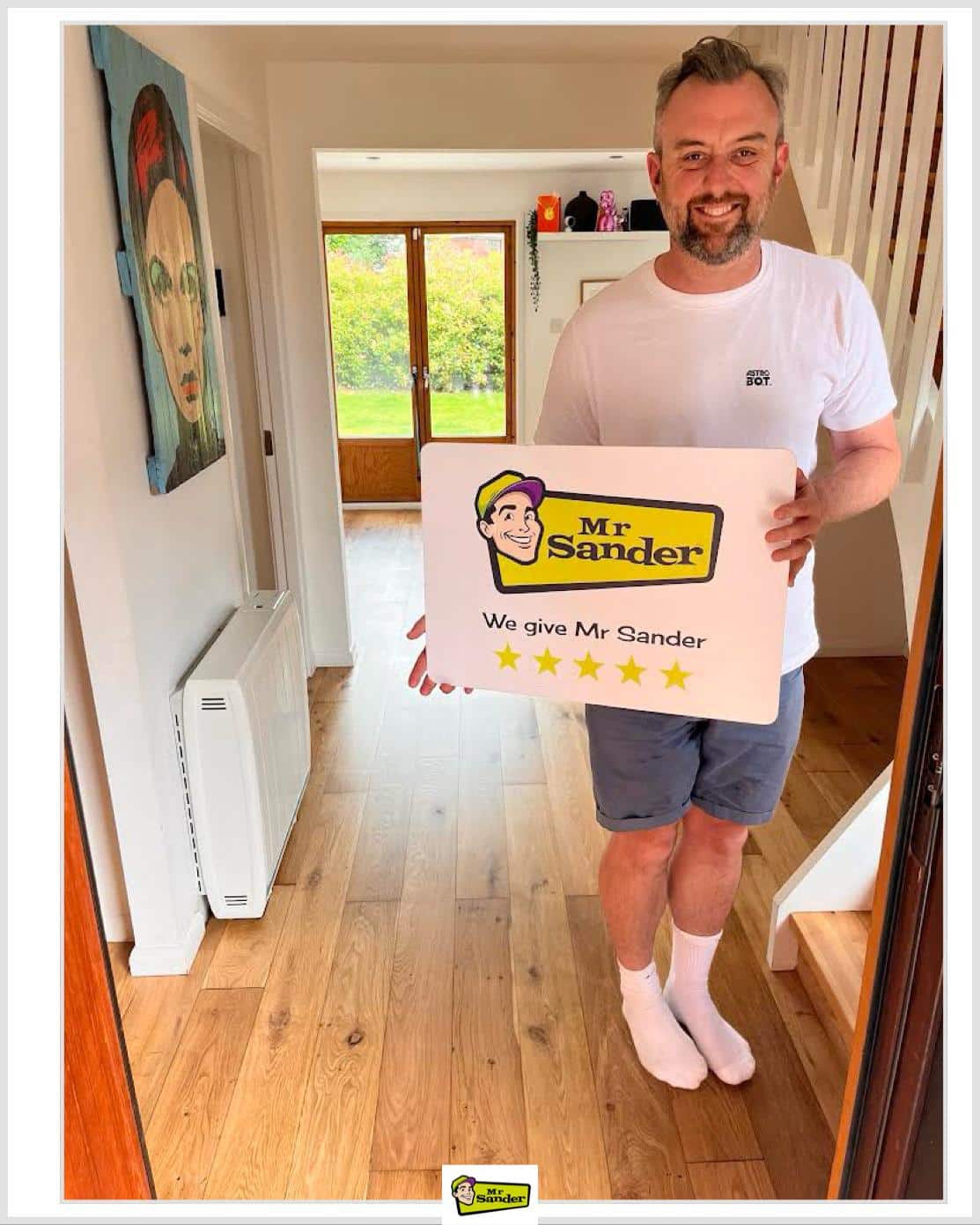 A satisfied customer holding a "We give Mr Sander 5 stars" sign in a beautifully refurbished hallway with sanded and refinished wooden floors by Mr Sander®. The hallway features a white wall with colorful artwork, a radiator, and glass doors leading to a lush garden in the background.