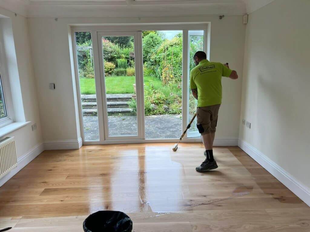 A worker wearing a green shirt and beige shorts is applying a finish to a newly sanded wooden floor using a large roller. The room has large glass doors that open to a garden with lush greenery outside. The left side of the floor is already finished and shiny, while the right side is in the process of being coated.