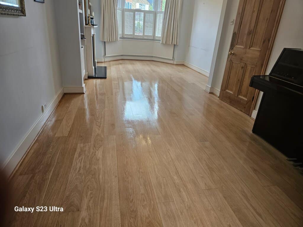 A beautifully refinished light oak hardwood floor in a spacious and well-lit living room. The floor has a glossy finish, reflecting natural light from a large bay window with white shutters. The room features minimal decor, with a wooden door and a piano adding a classic touch.
