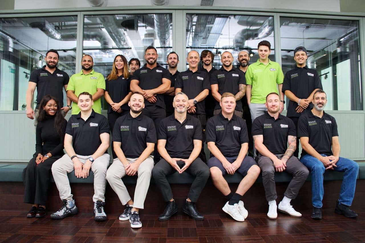 A group photo of the Mr Sander team, a floor sanding company in London and across the UK. The team members are dressed in black and green company-branded shirts, with a mix of men and women arranged in two rows: a seated row in front and a standing row behind. The photo is taken in a modern office with glass walls and visible air ducts, showcasing a professional and diverse team.