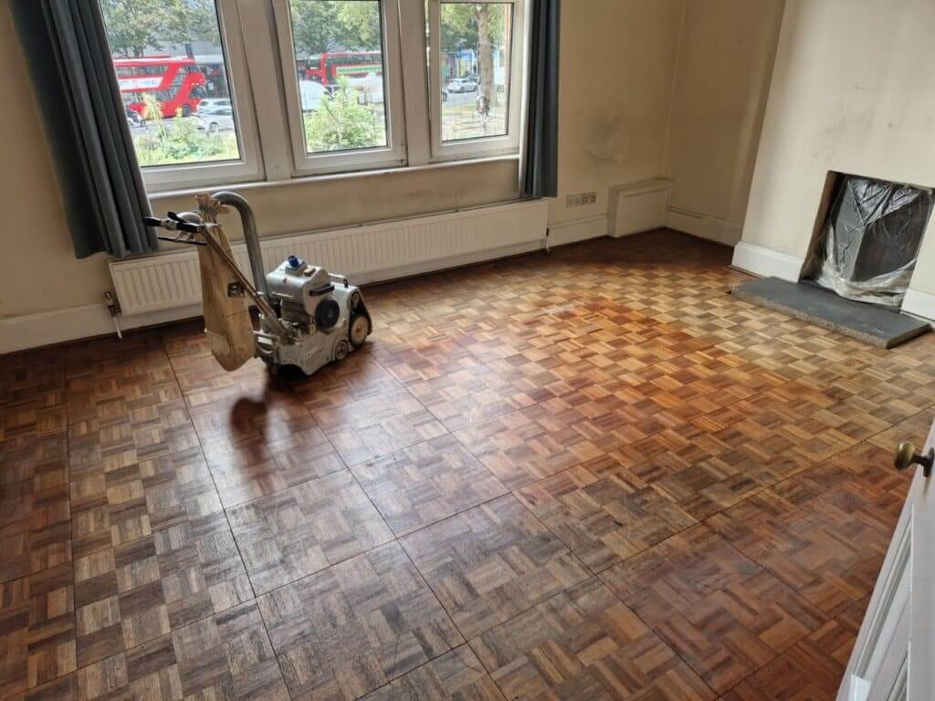 A professional from Mr Sander® sands a living room with dark-stained five fingers parquet wood floor in Milland, UK, using a Bona Belt (AMO110000.3) belt sander. The machine has a motor power of 2.2 kW, voltage of 230 V, frequency of 50 Hz, drum width of 200 & 250 mm, and a sanding belt size of 200 x 750 mm or 250 x 750 mm. The total weight of the machine is 74 kg.