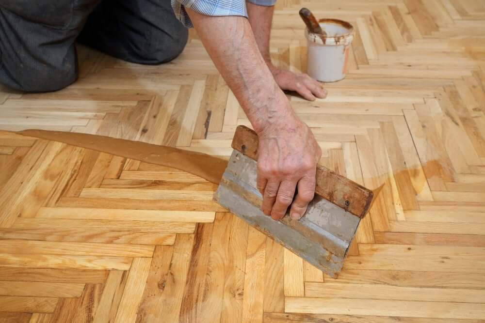 A professional applies a wood stain to a herringbone parquet floor using a scraper tool. A container of wood stain is visible in the background.
