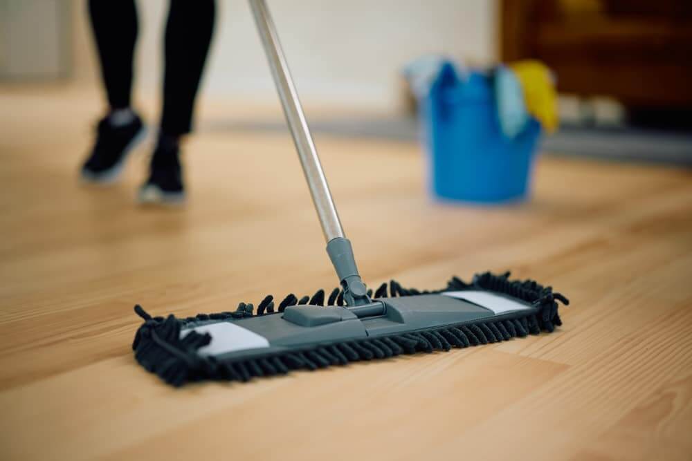 A person mops a freshly sanded and refinished light oak wood floor with a microfiber mop. A blue bucket with cleaning supplies is seen in the background, ensuring the floor remains clean and dust-free after the refinishing process.