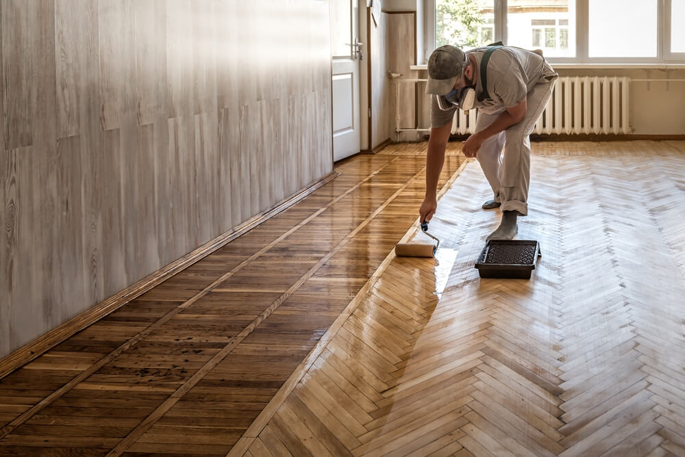 A professional floor refinisher wearing a protective mask applies a low-VOC, water-based finish to a herringbone hardwood floor using a paint roller and tray.
