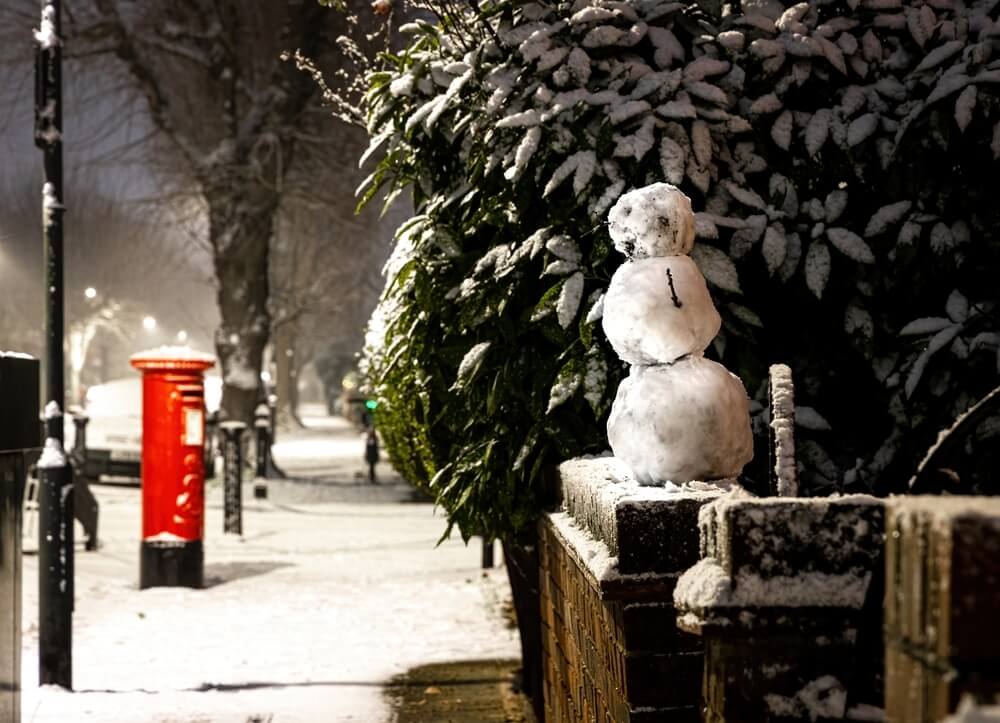 A nighttime winter scene in the UK, featuring a small snowman perched on a brick wall, with a classic red post box and snow-covered street in the background.