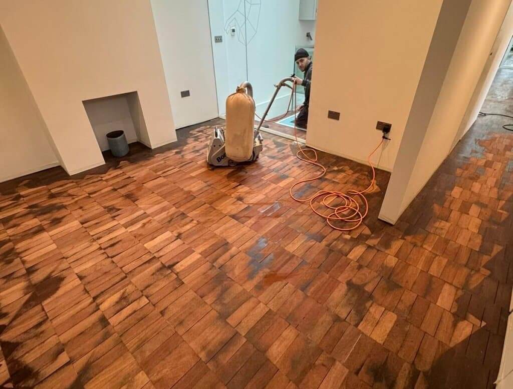 A worker from Mr. Sander® refinishes a unique wooden block parquet floor using a Bona sanding machine. The floor is mid-process, with darker and lighter sections showing the transition from sanding. The modern room features clean white walls and a minimalist design.