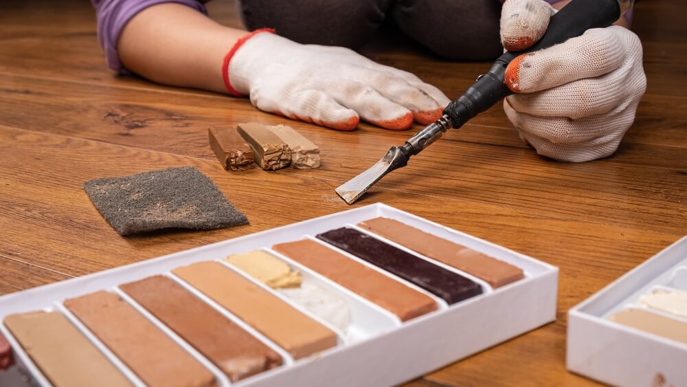Close-up of a floor repair process, showing hands in gloves using a tool and wax fillers on a wooden floor.
