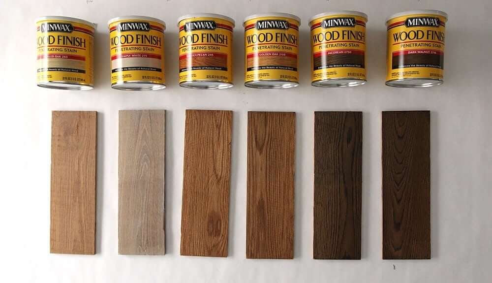 A selection of six Minwax Wood Finish penetrating stain cans is displayed above six wooden samples, each stained in different shades. The samples range from light to dark, showcasing the variety of finishes that can be achieved using the stains, including colours like Simply White, Golden Oak, Jacobean, and Dark Walnut.