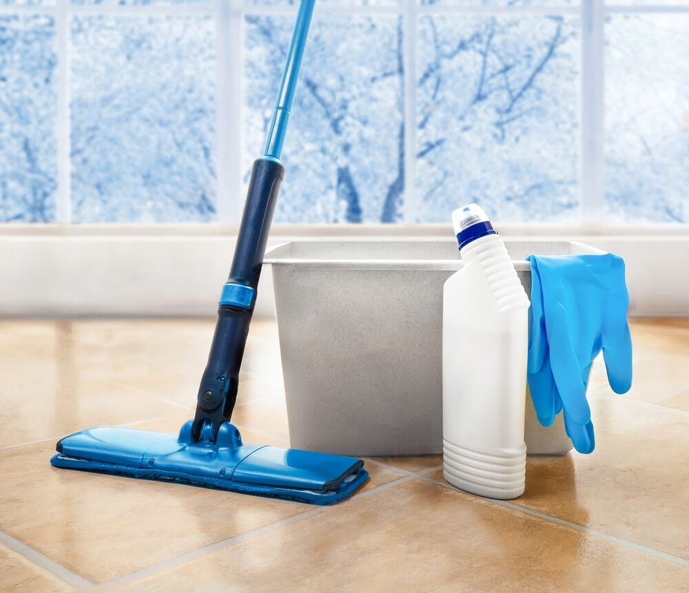 A tiled floor with cleaning tools, including a blue mop, a bucket, a bottle of cleaning solution, and blue rubber gloves. The background features a frosty window, suggesting a clean and fresh indoor environment. The image emphasizes post-refinishing care for maintaining flooring.