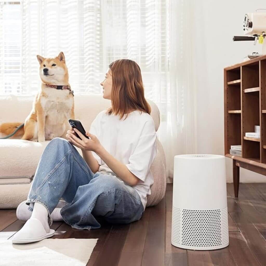  A cozy living room with a woman sitting on a polished hardwood floor, looking at her phone while her Shiba Inu dog sits on a nearby sofa. A white air purifier is placed on the floor, blending seamlessly with the modern decor. The floor has a rich, dark wooden finish that reflects the natural light streaming through sheer curtains.