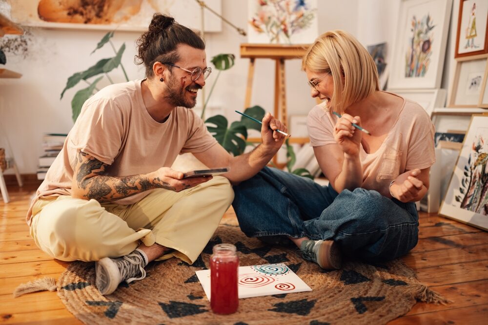 Two young artists sit cross-legged on a polished pine floor, laughing and painting an abstract canvas together in a bright, plant-filled studio.