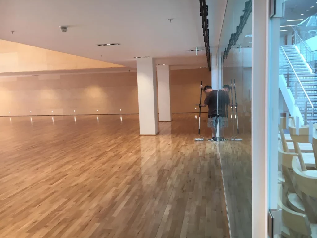 Technician applies a final, low-VOC lacquer with a wide applicator on a newly sanded maple strip floor inside a spacious school auditorium, mirrored in a floor-to-ceiling glass wall.