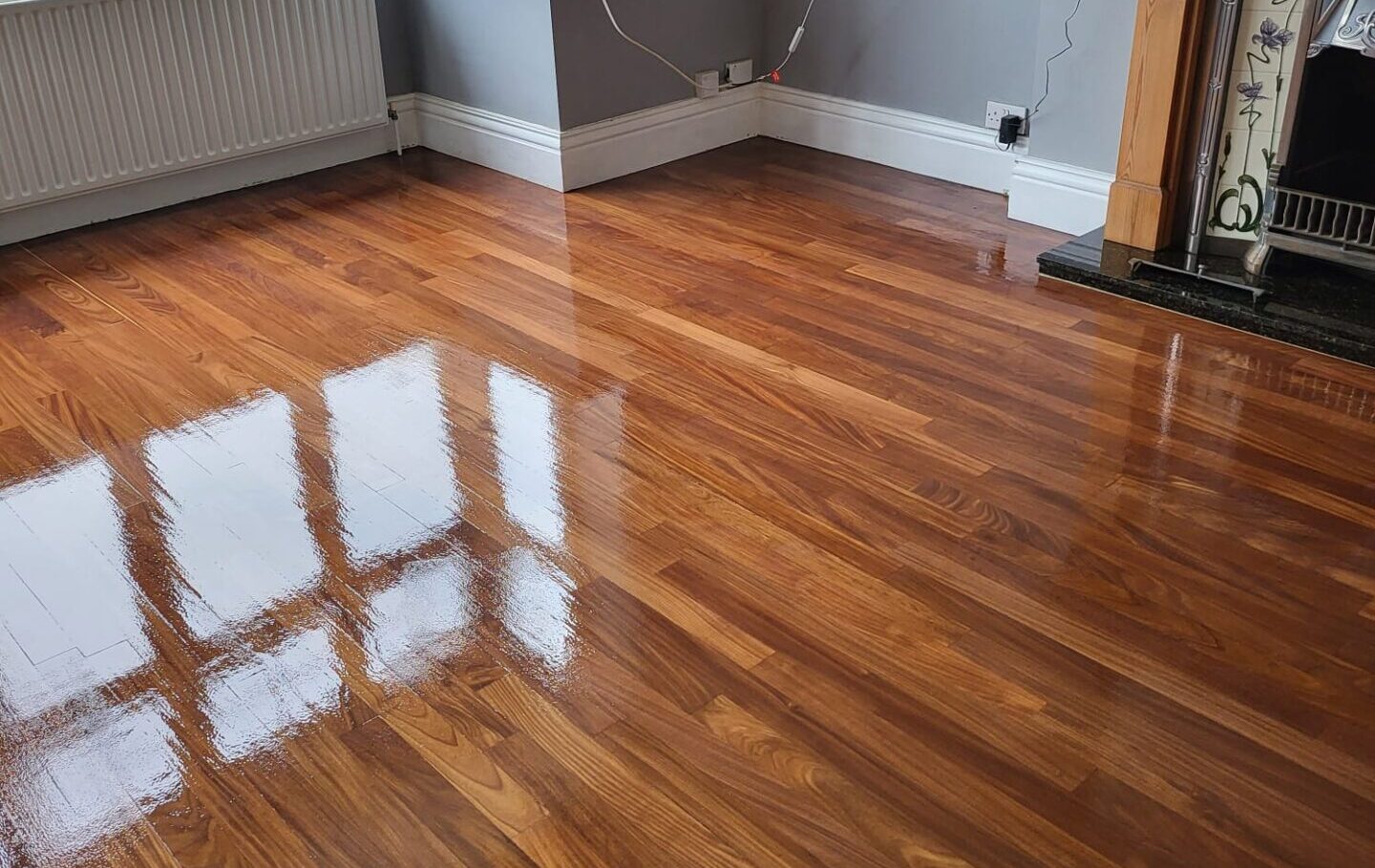 Bay-window living room with freshly refinished mahogany-toned hardwood flooring, high-gloss finish reflecting natural light beside a period fireplace.