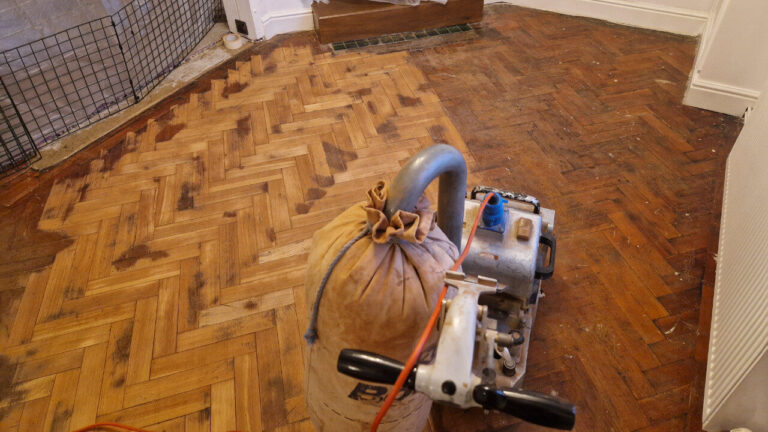Herringbone oak parquet wood block floor mid-sanding with a drum sander, dark coating being removed—on the way to beautifully restored parquet floors.