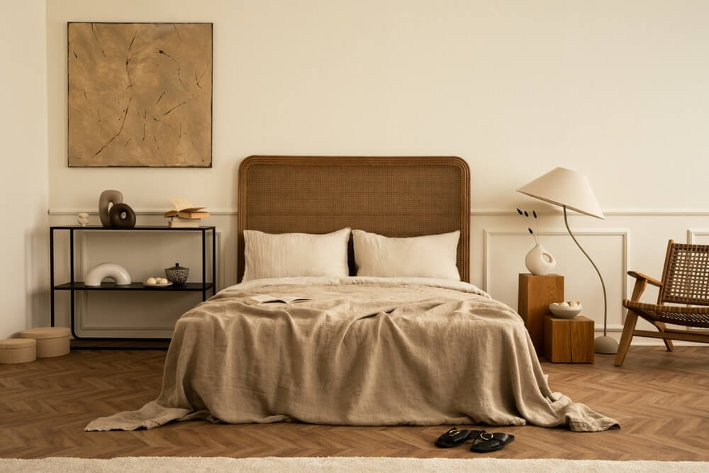 A cozy bedroom with a beautifully refinished parquet wood floor, featuring a warm brown tone. The room is decorated with a woven headboard, beige bedding, minimalist furniture, and natural textures like a wooden chair and side table. A piece of abstract art hangs on the wall, complementing the neutral color scheme.