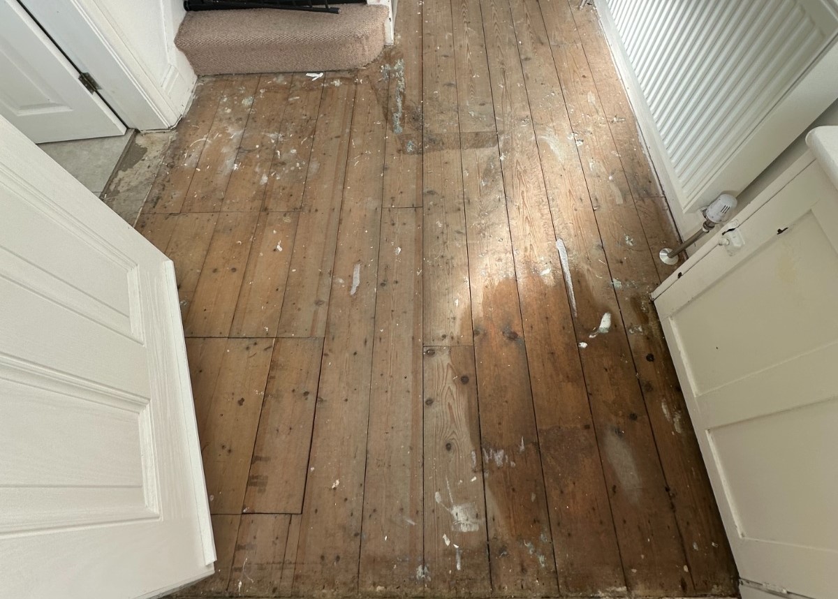 A top-down view of worn pine softwood floorboards covered in paint stains and old varnish before the refinishing process begins.