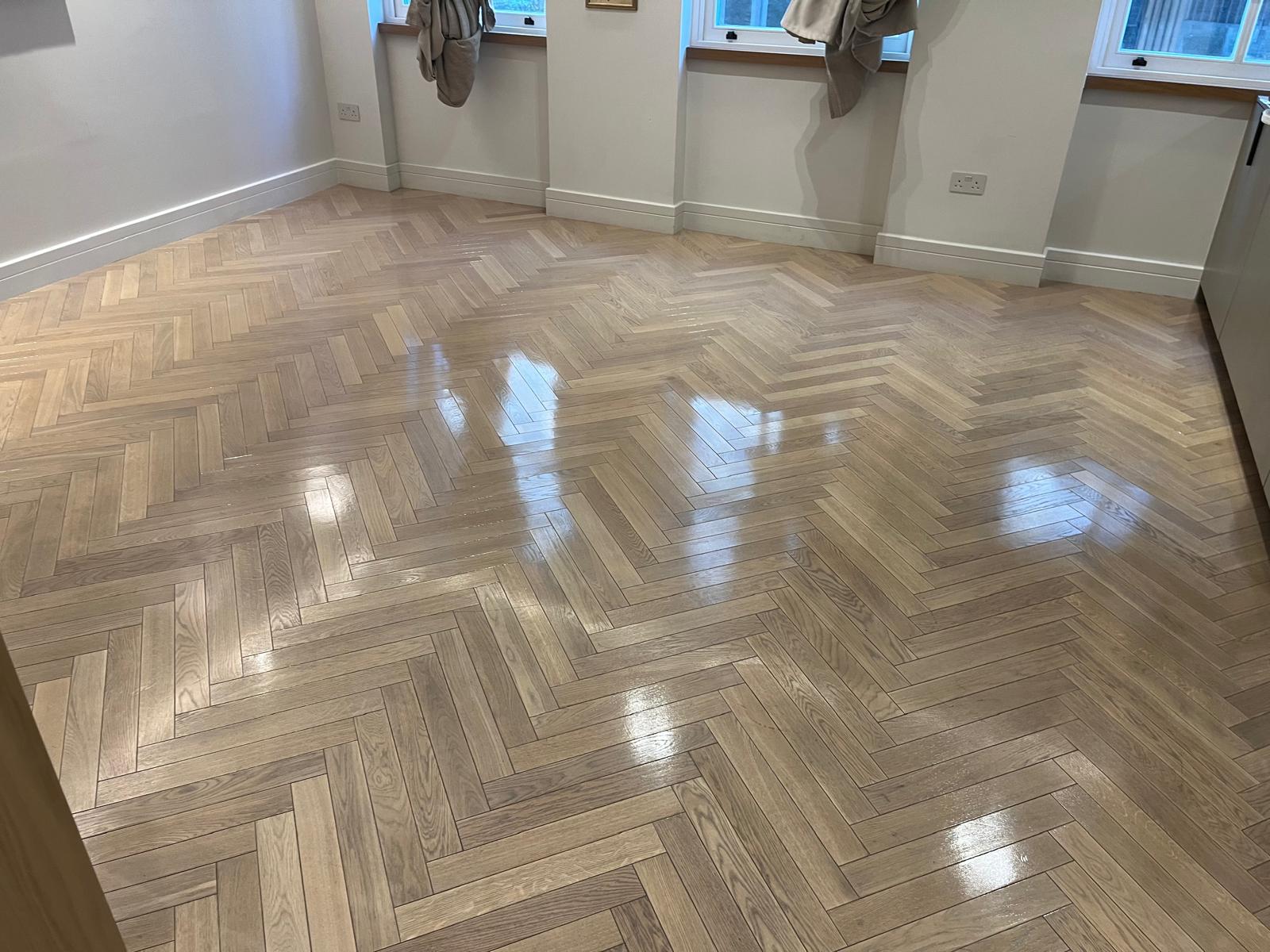 A high-gloss, light oak herringbone parquet floor in a bright room, showcasing the best finish for reflecting natural light and highlighting wood grain.