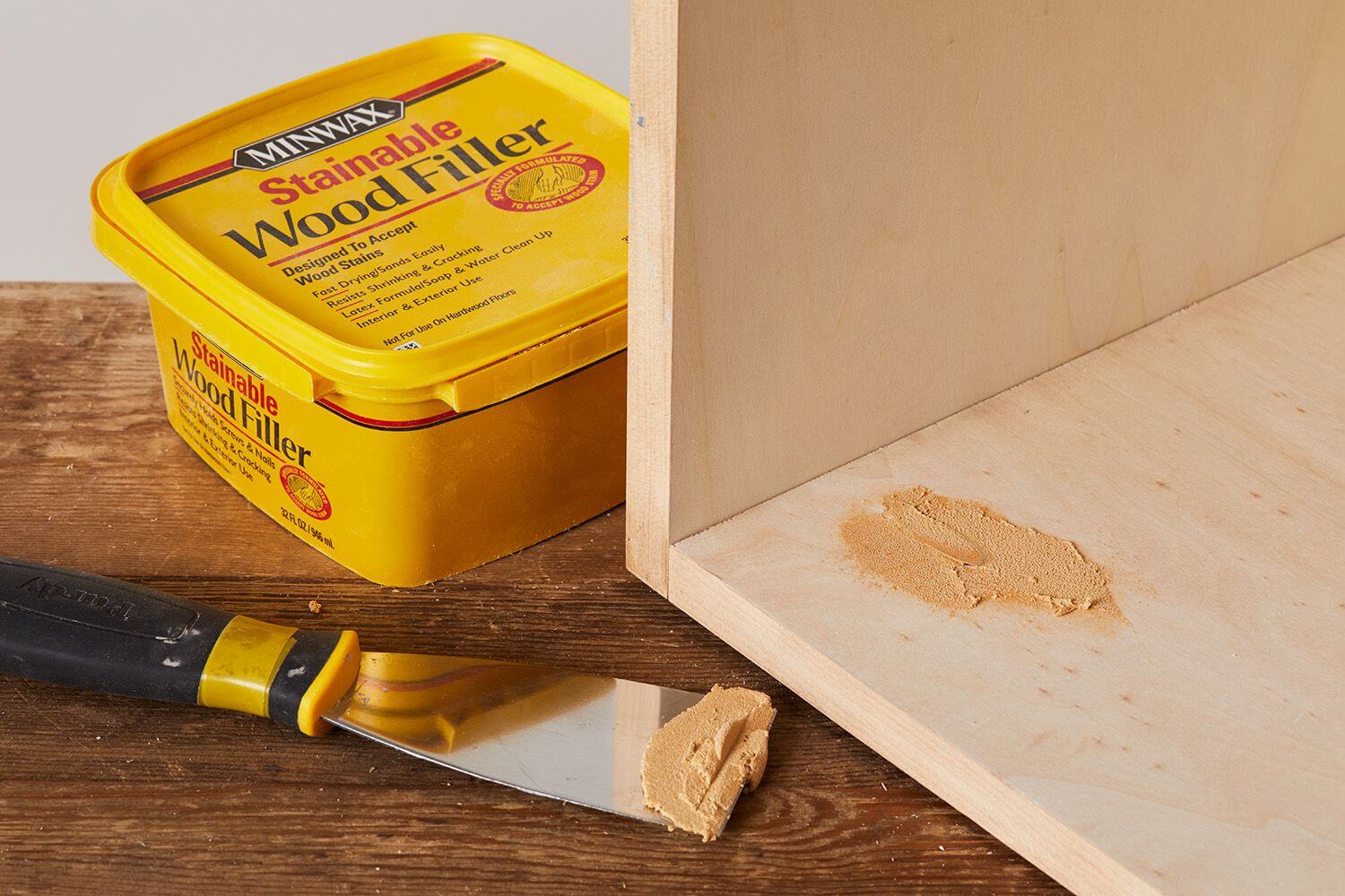 A container of Minwax Stainable Wood Filler with a putty knife and some applied wood filler on a wooden surface, demonstrating its use in filling gaps or imperfections in wood.