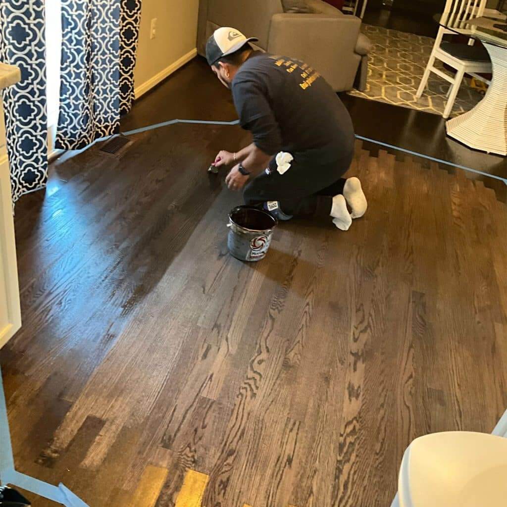 A professional from Mr Sander® applying a dark stain to a sanded wooden floor in a living room. The worker is using a brush and a bucket of stain, carefully covering the floor to achieve a uniform finish.