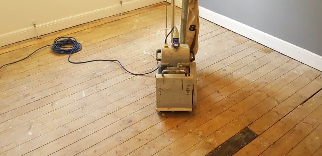 A professional from Mr Sander® is sanding a pine floor in a living room using a Bona Belt sander (AMO110000.3). The machine features a 2.2 kW motor, 230 V voltage, 50 Hz frequency, and a sanding belt size of 200 x 750 mm or 250 x 750 mm. The total weight is 74 kg.