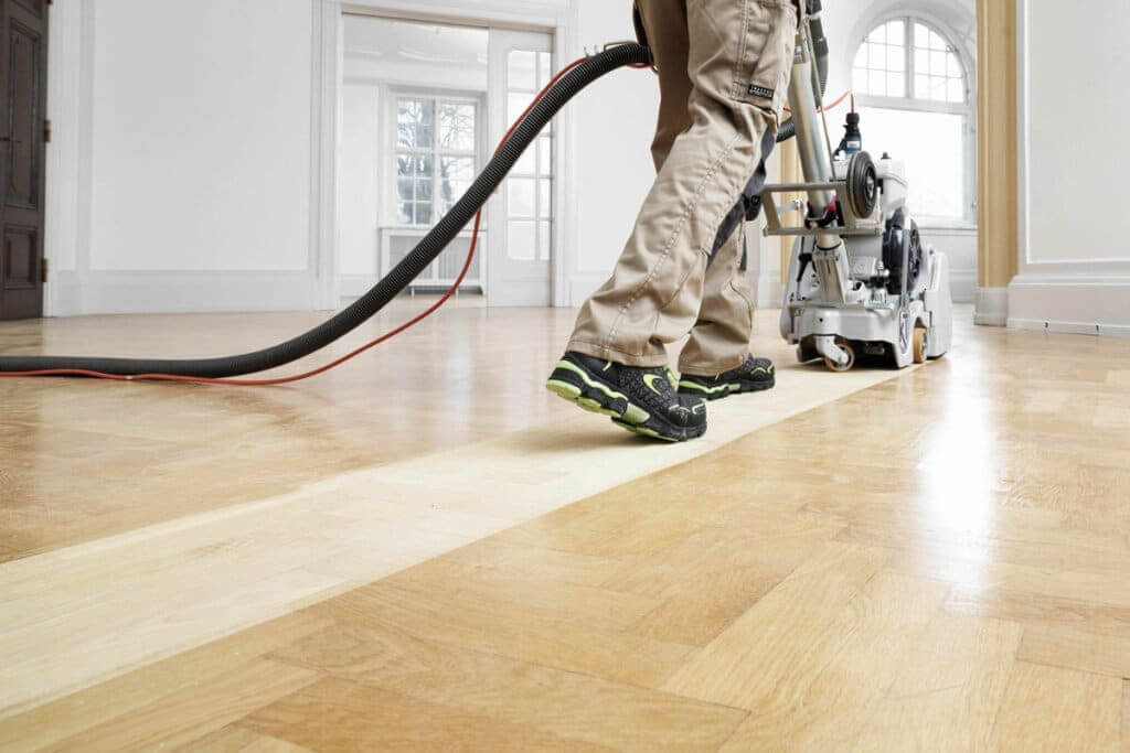 A man from Mr Sander® sanding a hardwood floor in a living room in Oxshott , UK, using the Bona Belt UX sander (AMK173100). The Bona Belt UX features a 2.2 kW motor, operates at 230 V and 50 Hz, with a sanding belt size of 200x750 mm, drum width of 200 mm, and weighs 85 kg.