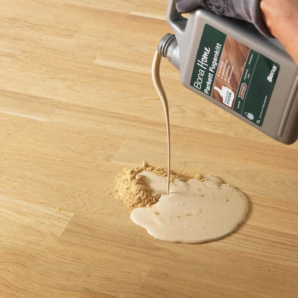 A close-up of a person pouring Bona Home wood floor filler onto a light-colored wooden floor. The filler is being mixed with sawdust to match the floor color, indicating a step in floor refinishing.
