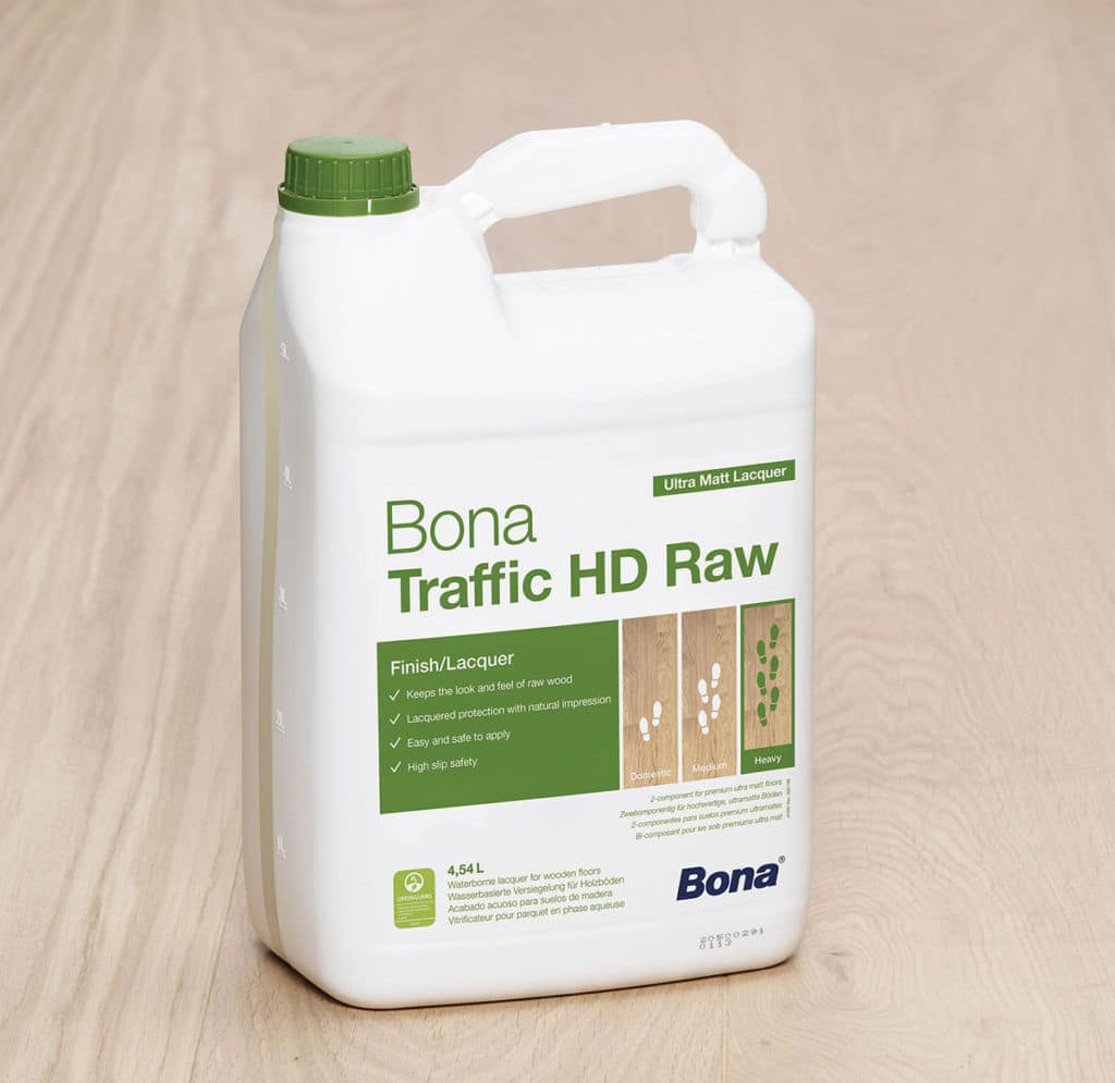 A white container of Bona Traffic HD Raw, an ultra-matt lacquer, is displayed on a wooden floor. The label highlights its features, including maintaining the look and feel of raw wood, providing lacquered protection with a natural impression, being easy and safe to apply, and offering high slip safety. The container has a green cap and a handle for easy carrying.