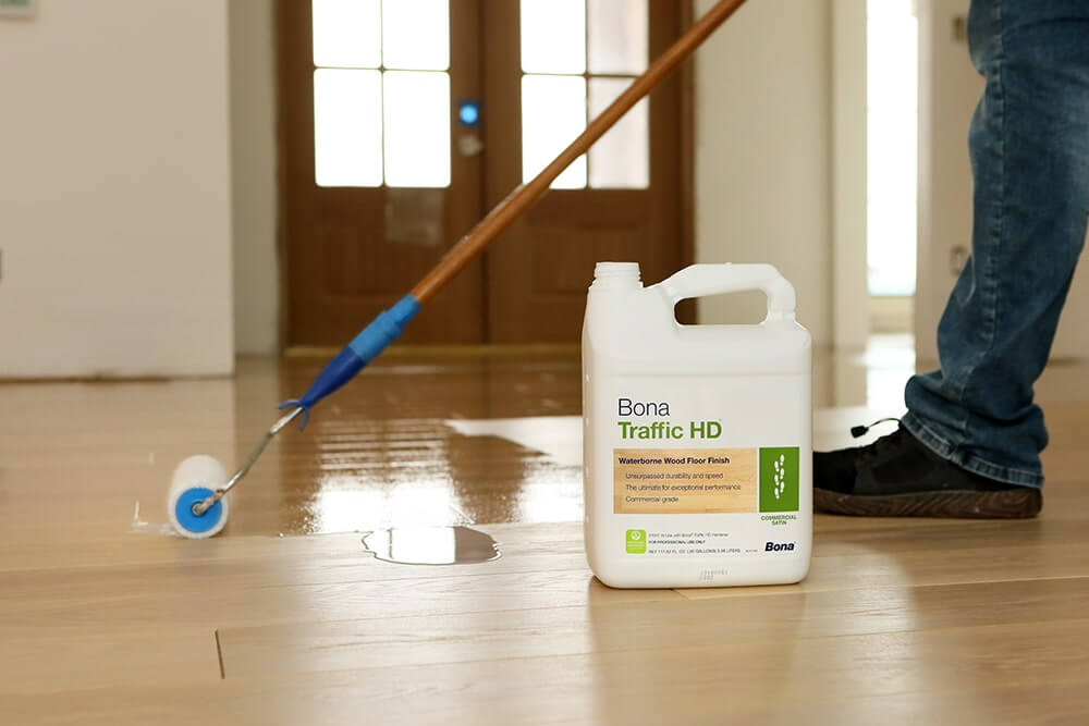 A man from Mr Sander® is applying a waterborne wood floor finish using a roller in a living room. The floor finish being used is Bona Traffic HD, as shown by the container placed nearby. The floor appears glossy and well-refinished, and the background shows a wooden door with glass panels.