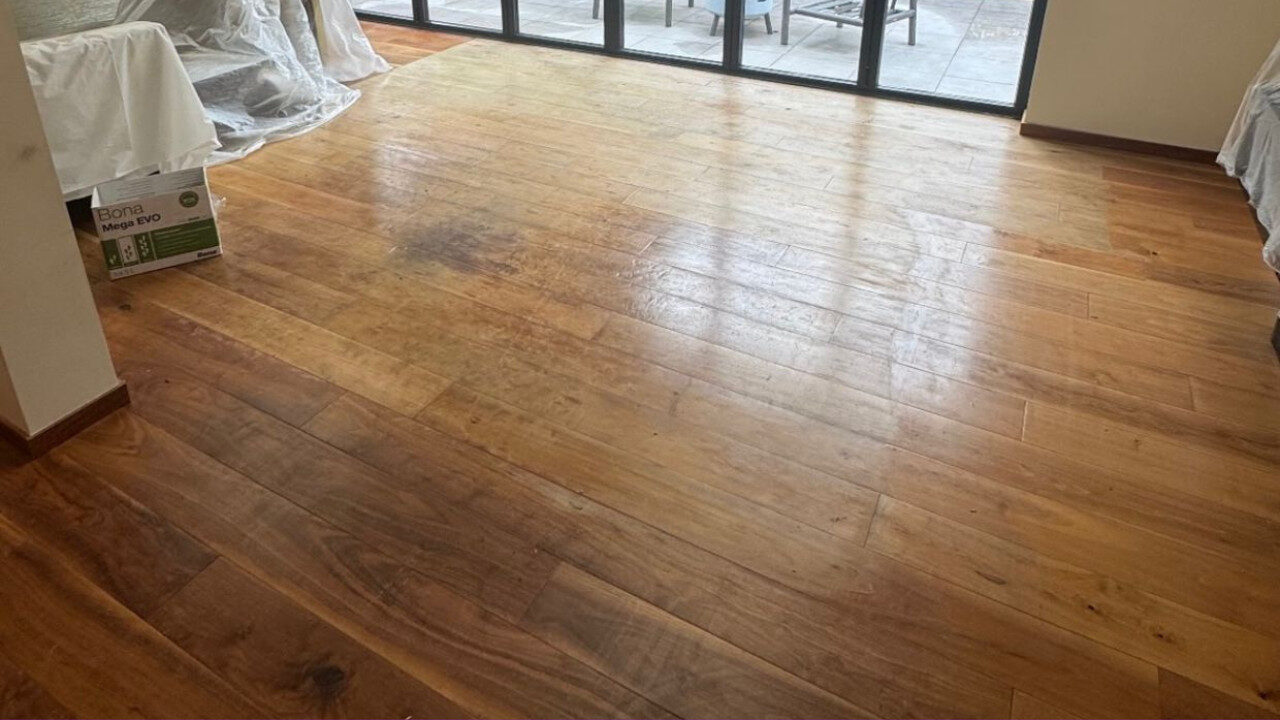 A wide-angle view of an engineered oak wide plank floor before restoration, showing natural wear and varying honey-brown wood tones near a large glass door.
