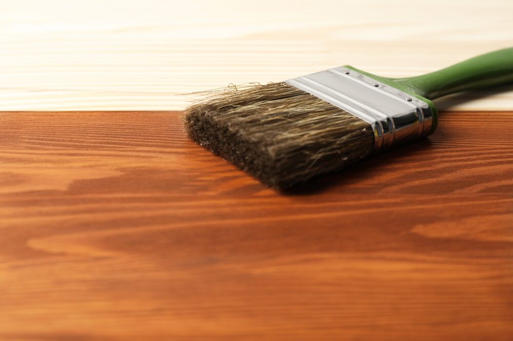 Close‑up of a paintbrush sweeping rich Wood Stain across pale pine boards, revealing warm grain patterns.