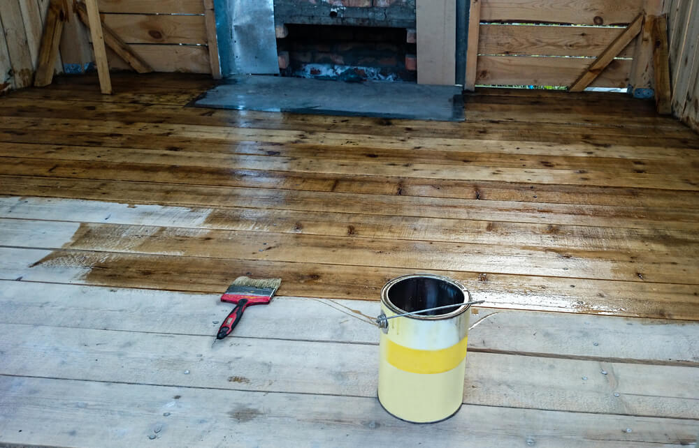 A partially refinished wooden floor with a distinct line separating the stained and unstained sections. A paintbrush with a red handle and a partially used can of stain are placed on the unstained portion of the floor. The background shows a rustic wooden wall and a partially visible fireplace, adding to the room's unfinished look.