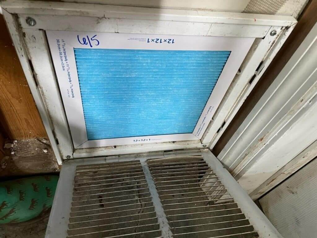 A close-up view of an air vent or HVAC filter slot with a clean blue filter installed. The frame of the vent appears slightly aged, with markings indicating "6/5." The surrounding area shows minor dust and wear, suggesting the filter has been recently replaced or serviced.