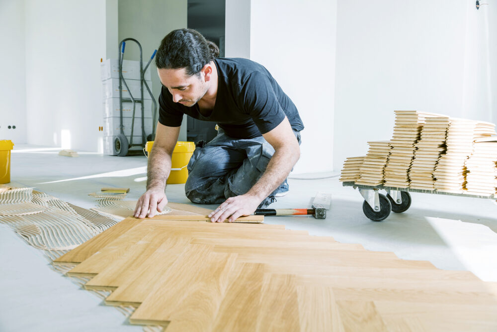 Professional fitter aligning engineered oak herringbone parquet during a glue-down installation — Chevron vs Herringbone comparison showing classic herringbone pattern.