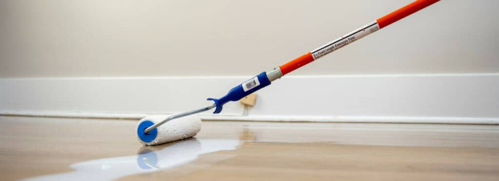 Application of finish on a hardwood floor using a paint roller with an orange handle.