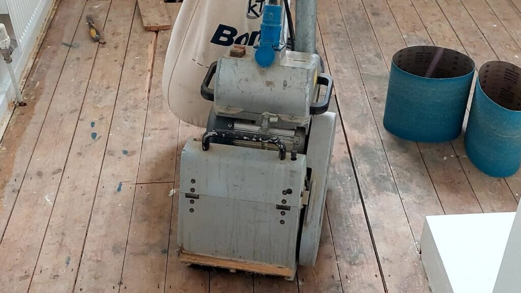 A man from Mr Sander® is sanding a pine floor in the living area of a home in, UK, using a Bona Belt (AMO110000.3) sanding machine. The Bona Belt features a 2.2 kW motor, 230 V voltage, 50 Hz frequency, 200 & 250 mm drum width, and a sanding belt size of 200 x 750 mm or 250 x 750 mm, with a total weight of 74 kg.