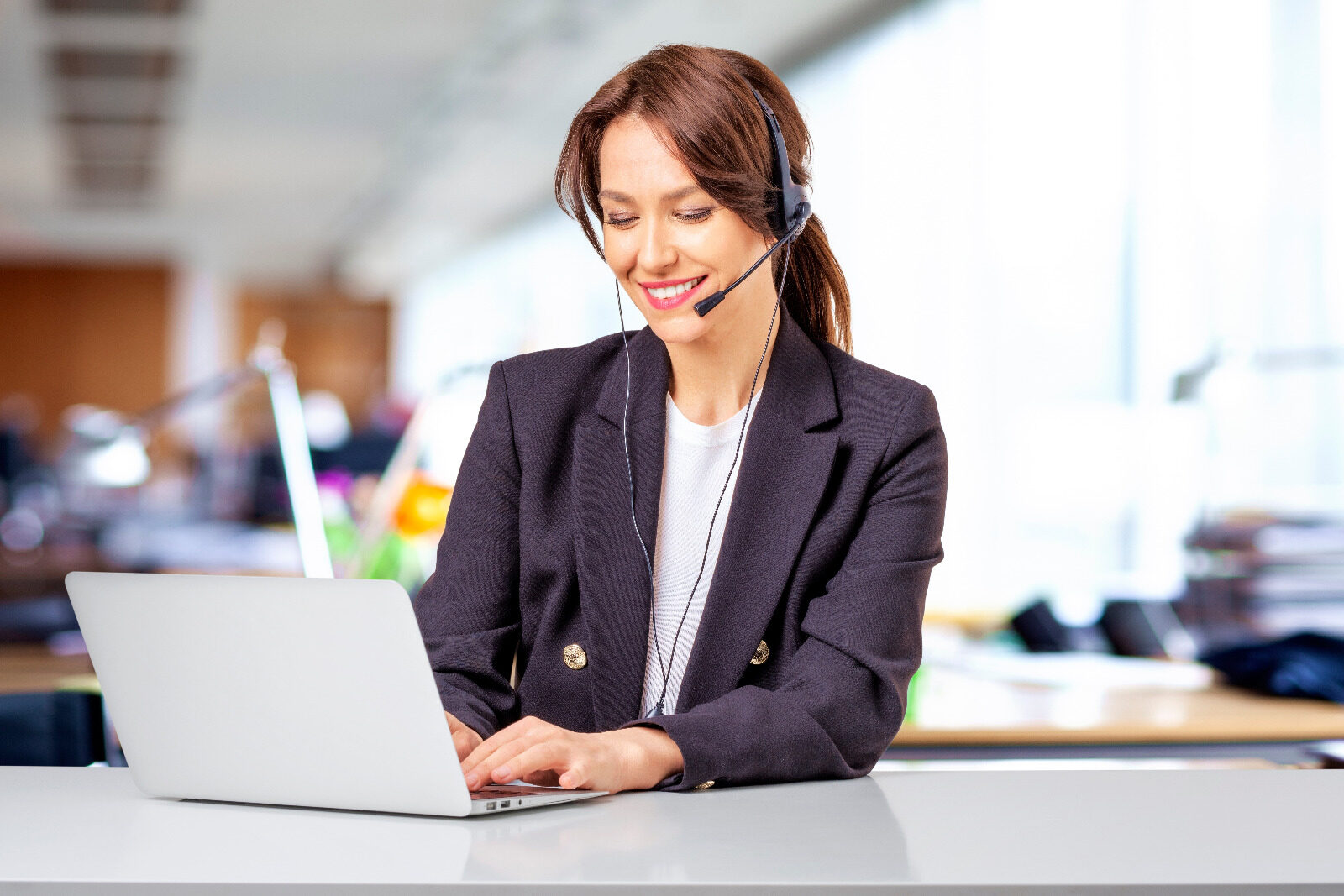 Smiling customer support advisor wearing a headset and typing on a laptop in a bright office