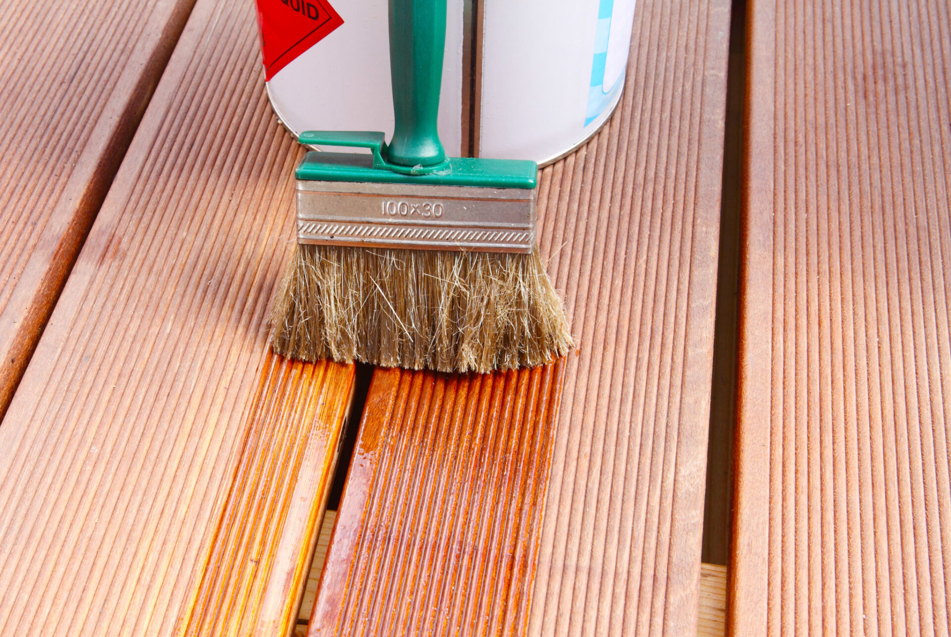 Brush and tin of stain on grooved softwood timber decking floor, showing Deck Stain Colour Ideas with a fresh tinted finish being applied to the wooden boards.