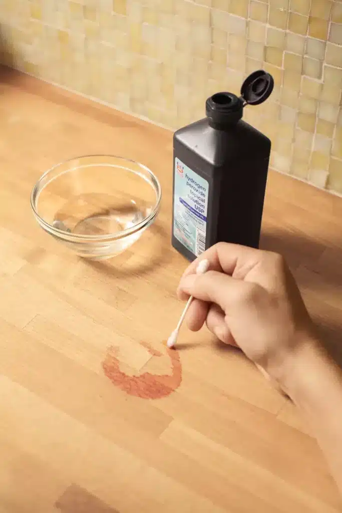 Hand using a cotton swab to clean a wine stain on a wooden countertop with hydrogen peroxide.
