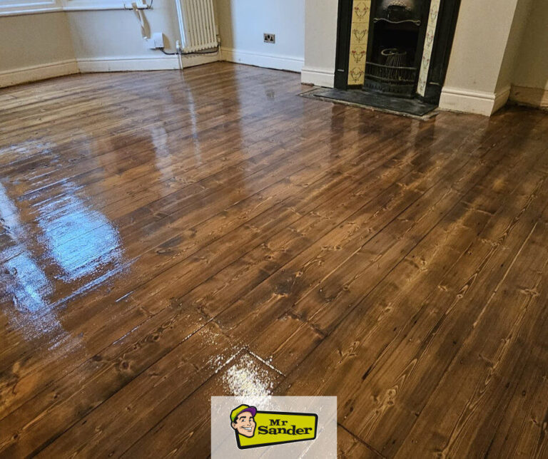 Freshly finished pine floorboards with a glossy coat after DIY sanding in a living room; reflection near bay window and Victorian fireplace — Mr Sander®.